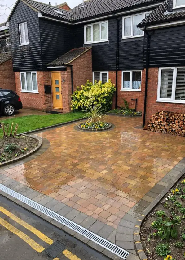 Wet block-paved driveway with central flower bed, adjacent garden, and a brick and black-clad UK house with stacked logs.