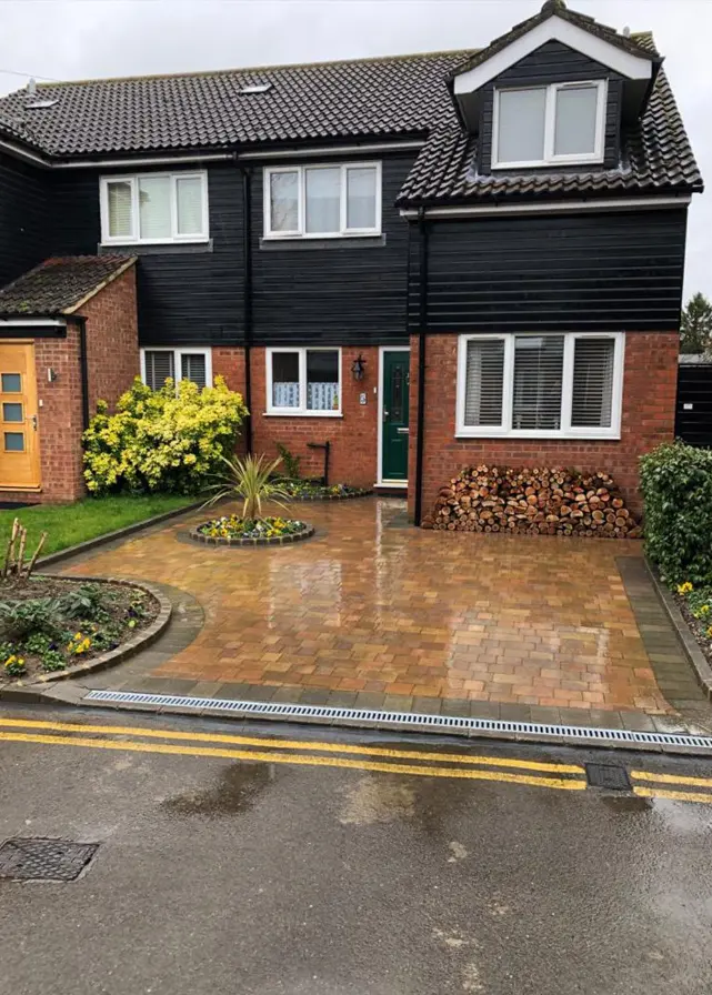 British semi-detached house with black cladding, green door, paved driveway, circular flower bed, and stacked firewood.