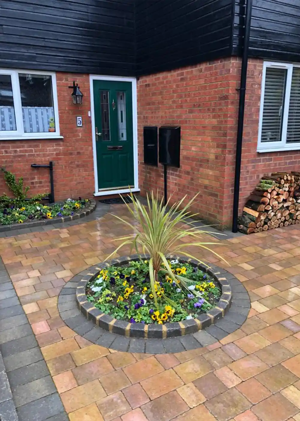 Front entrance of a modern British brick house with a green door, two black mailboxes, white-framed windows, neatly stacked firewood, and a circular flower bed with colourful pansies and a central spiky plant, all set on a paved driveway.