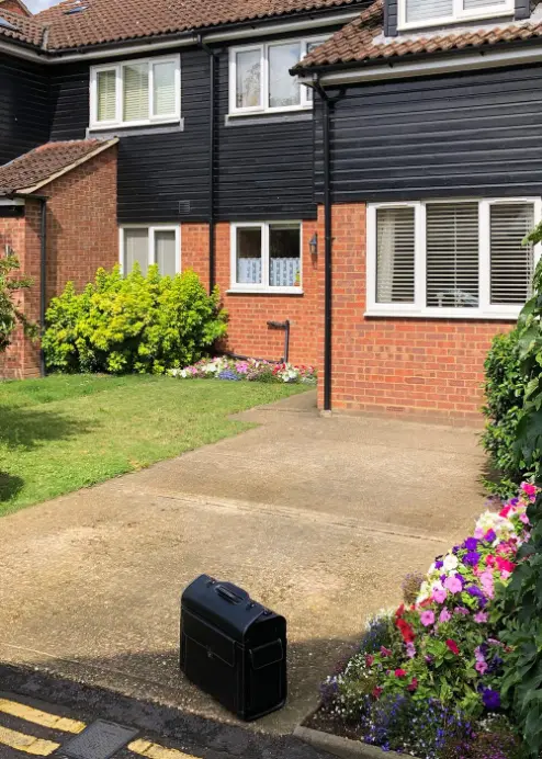 Driveway of a UK semi-detached house with brick and black cladding, featuring flowers and a briefcase on the concrete driveway.