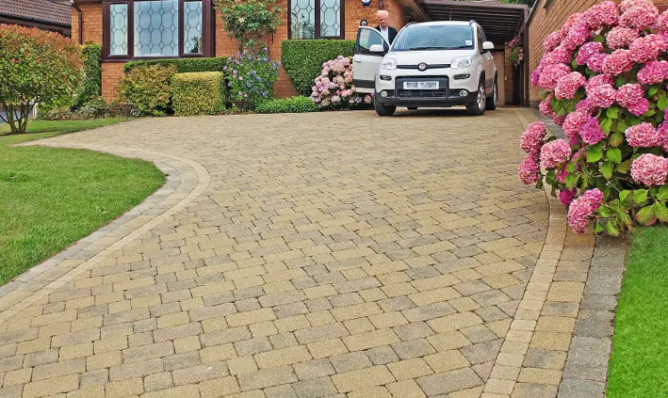 Curved driveway with a man stepping out of a white car near a brick house, pink flowers and a green lawn on sides.