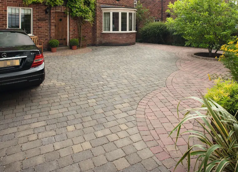 Black Mercedes parked on gray and beige circular cobblestone driveway with red brick house, wooden door, potted plants, curved flower bed, and green shrubs under cloudy sky.