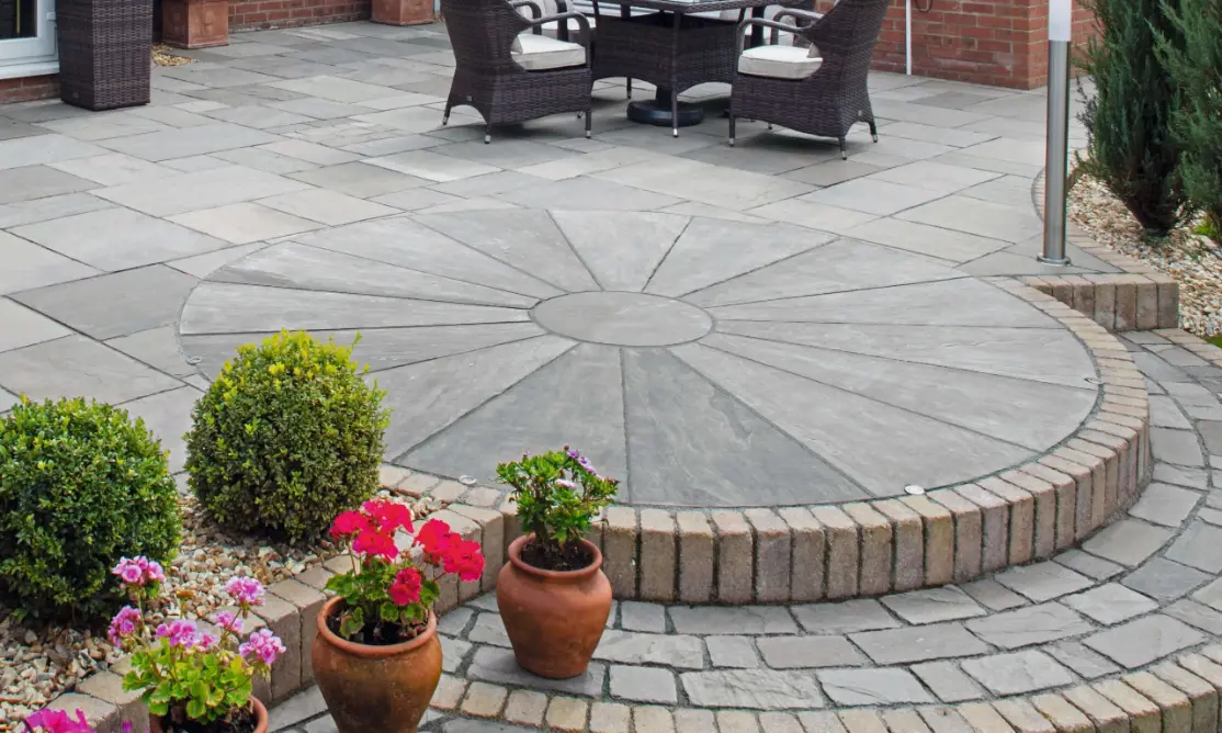 Outdoor patio with circular stone feature surrounded by brick edging, potted flowers, and wicker chairs with cushions.
