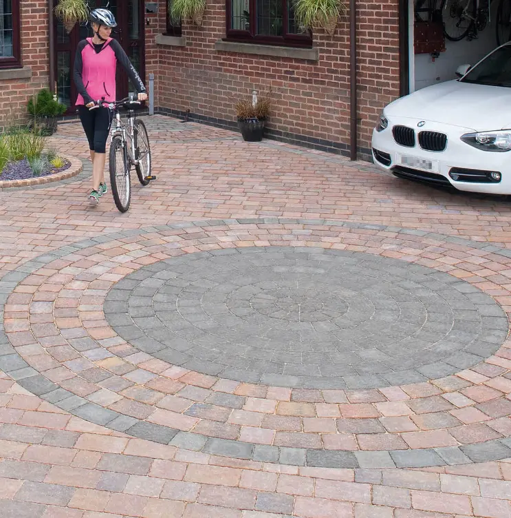 Woman in cycling helmet walking bicycle on circular-patterned block paved driveway beside parked white car.
