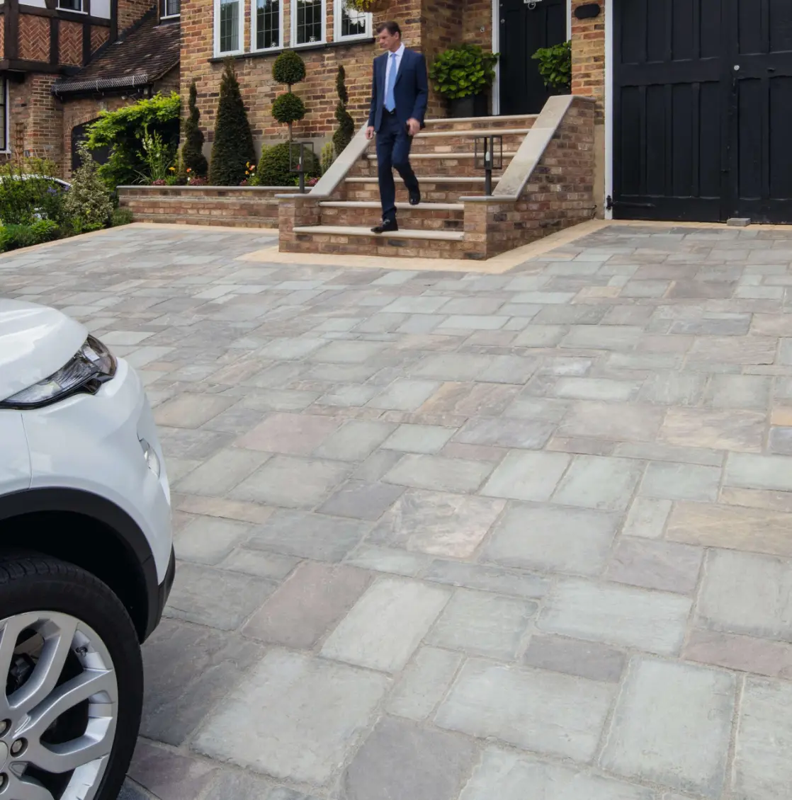 Man in blue suit walking down brick steps from house onto gray and tan stone driveway with white car parked on the left.