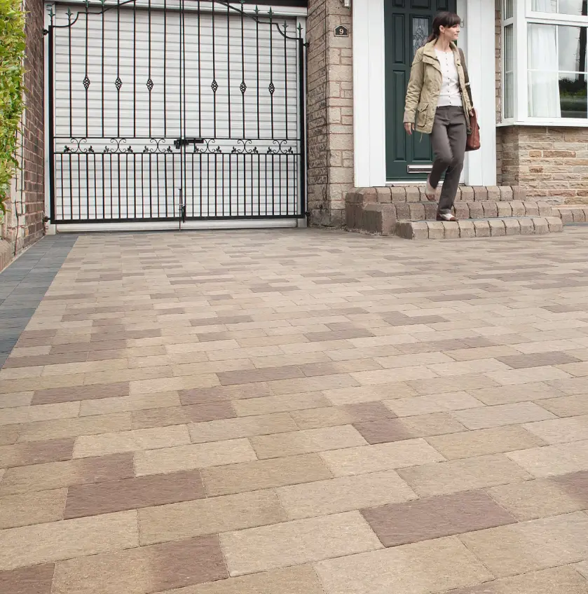 Woman walking down brick steps from a house onto a driveway with beige and brown paving stones and a black metal gate.