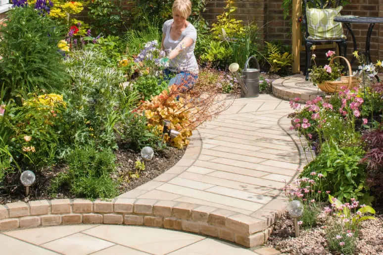 Woman tending to a garden bed filled with colorful flowers and plants along a curved beige stone walkway bordered by low brick edging, with a metal watering can and black metal table with chair and cushion in the background.