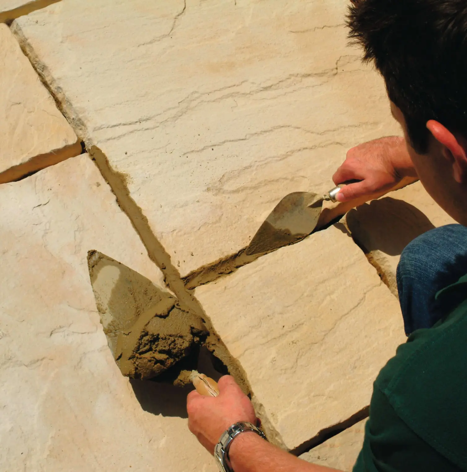 Man using trowels to fill and smooth mortar between large sandstone paving slabs during outdoor stonework installation.