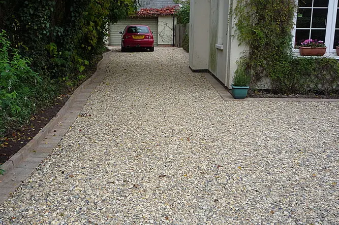 Gravel driveway bordered with bricks, leading to a red car parked near a garage, with green plants and shrubs along the left side and climbing ivy on the right side of a house.