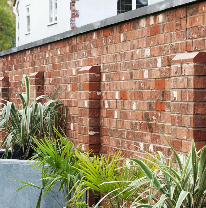 Red brick garden wall with decorative pillars, large grey planter, and variegated green plants in front.