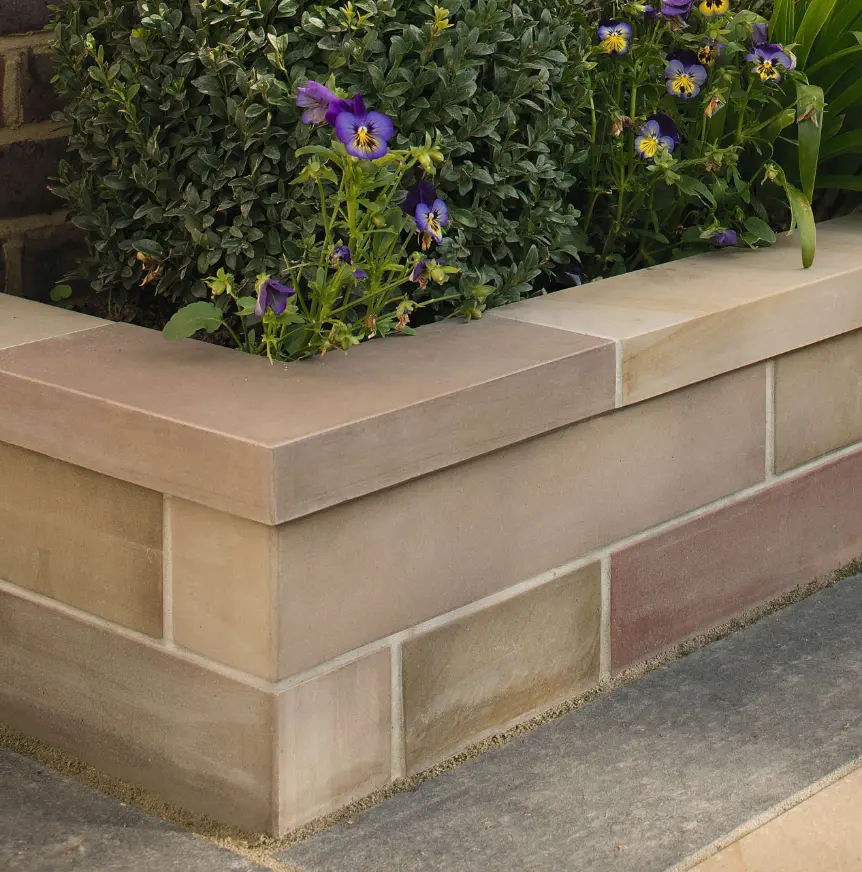 Corner of beige stone raised garden bed with purple pansies and green foliage growing inside on paved garden path.