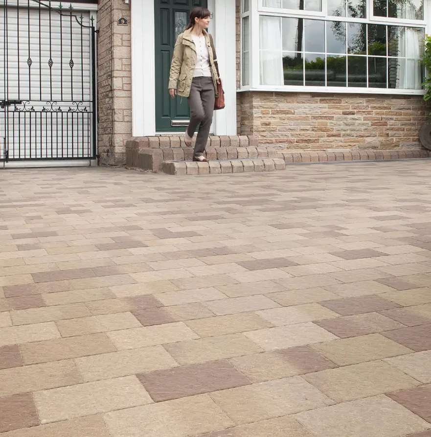 Woman walking down brick steps to a patterned paved driveway outside a house with stone and brick exterior.